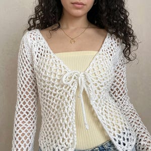 May include: A white, open-weave crochet cardigan with long sleeves and a tie closure at the chest. The cardigan is worn over a yellow top. The crochet pattern creates a net-like appearance. The style is casual and bohemian.