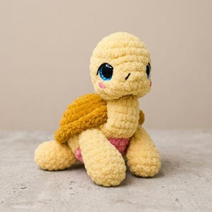 Könnte beinhalten: Ein gehäkeltes Schildkröten-Stofftier mit hellgelbem Kopf und Gliedmaßen, einem strukturierten senfgelben Panzer und einem rosafarbenen Bauch. Die Schildkröte hat große, glänzende blaue Augen mit schwarzen Pupillen und kleine rosa Wangen.
