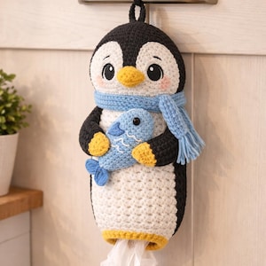 Könnte beinhalten: Gehäkelter Pinguin-Taschentuchhalter in Schwarz, Weiß, Gelb und Blau. Der Pinguin hat einen blauen Schal und hält einen blauen Fisch. Der Taschentuchhalter hängt an einem Schrankgriff und gibt Taschentücher von unten aus.