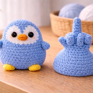 Puede incluir: Dos artículos de ganchillo hechos a mano: un pingüino azul con detalles blancos y pies amarillos, y una mano azul haciendo un gesto grosero. El pingüino tiene ojos negros y un pico naranja. Están sobre una superficie de madera.