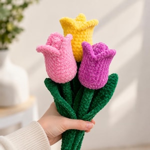 Puede incluir: Un ramo de tres tulipanes de ganchillo hechos con hilo suave y afelpado en colores amarillo vibrante, rosa claro y morado. Cada flor está unida a un tallo verde largo y texturizado con hojas verdes a juego, tejido con puntos consistentes.