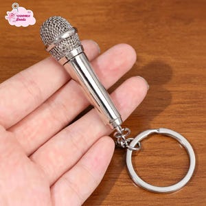May include: A silver-colored miniature microphone keychain. The microphone has a mesh head and a cylindrical body, attached to a keyring. The keychain is held in a hand against a wooden background. The keychain is a novelty item.