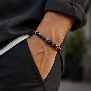 Op de afbeelding: Een kralenarmband met een reeks gladde, ronde stenen in tinten dieprood, donkerblauw en bruin, geaccentueerd door twee kleine, gedetailleerde zilverkleurige metalen tussenstukjes. De kralen zijn geregen aan een elastisch koord voor een flexibele pasvorm.