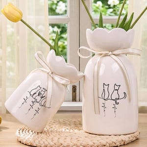 May include: Two white ceramic vases with yellow tulips. The vases have a unique shape, with a scalloped top and a ribbon tied around the neck. One vase has a cat illustration and the text "Sofia". The other vase has a cat illustration and the text "Sofia". The text "Custom vases" is at the top.