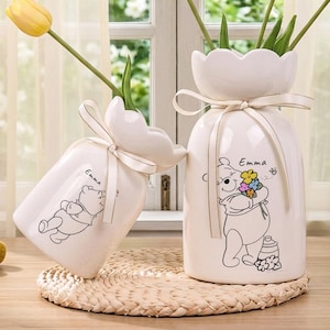 May include: Two white ceramic vases, each with a Winnie the Pooh illustration and the name "Emma". They hold yellow tulips. The vases are tied with white ribbons and sit on a woven mat. The text "Custom vases" is at the top.