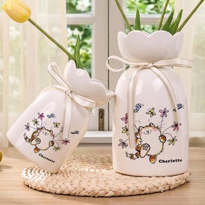 Op de afbeelding: Twee witte keramische vazen met een kat- en vlinderontwerp, met gele tulpen. De vazen hebben een lint om de hals en de tekst "Charlotte" onderaan. De tekst "Custom vases" staat bovenaan.