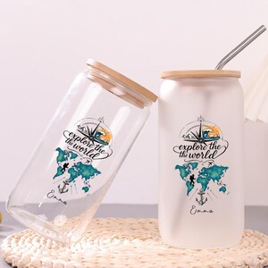 May include: Two glass tumblers with bamboo lids and metal straws. One is clear, the other frosted. Both feature a graphic of a world map, a compass, and the text "explore the world" with the name "Emma" printed in black script below the map.