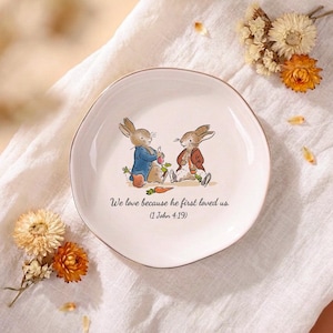 Può includere: Un piattino rotondo in ceramica color bianco sporco con l'illustrazione di due coniglietti che indossano giacche. Il testo recita: "We love because he first loved us. (1 John 4:19)." Il piatto ha un bordo liscio leggermente irregolare e finitura lucida.
