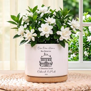 May include: A white ceramic planter with a wooden base, filled with a flowering plant with white blossoms and green leaves. The planter has text that reads "Our New Home, New Memories, 12 Chocolate Street, Deborah & Mick, 2023".