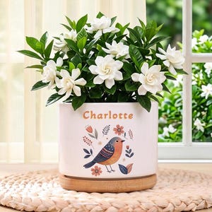 Op de afbeelding: Een witte cilindervormige keramische plantenpot met een houten voet, voorzien van de naam Charlotte in oranje tekst boven een kleurrijke illustratie van een vogel omringd door bloemmotieven, met een groene gardenia met witte bloemen.