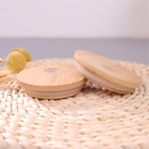 May include: Two circular bamboo lids for drinkware, each featuring a smooth, light-toned wood grain finish and a central circular opening. A translucent silicone gasket is fitted around the underside edge of each lid to ensure a secure seal.