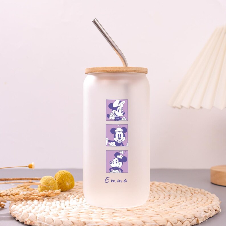 Puede incluir: Un vaso de vidrio esmerilado con tapa de bamb&uacute; y pajita de metal. El vaso presenta tres gr&aacute;ficos cuadrados morados apilados de un personaje de rat&oacute;n de dibujos animados y el nombre Emma impreso en una fuente cursiva morada.