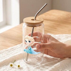 May include: A clear glass tumbler with a bamboo lid and a bent metal straw. The glass features a cartoon illustration of a blonde character in a blue dress with snowflake accents. The design includes small blue snowflakes scattered around the figure.