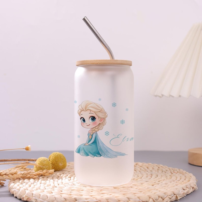 May include: A frosted glass tumbler with a bamboo lid and a bent metal straw. The tumbler features a cartoon illustration of a blonde character in a blue dress with the name Elsa written in cursive script and small blue snowflake accents.