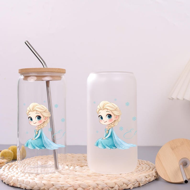 May include: Two glass tumblers with a cartoon illustration of a blonde character in a blue dress surrounded by small blue snowflakes. One tumbler is clear glass with a bamboo lid and metal straw, while the other is a frosted glass tumbler.