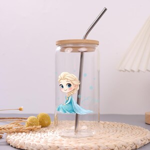 May include: A clear glass tumbler with a bamboo lid and a metal straw. The glass features a cartoon illustration of a blonde character in a light blue dress with a long braid, surrounded by small blue snowflake icons and the name Elsa in script.