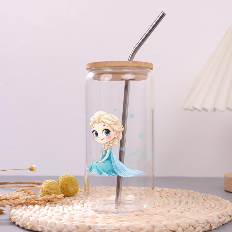 May include: A clear glass tumbler with a bamboo lid and a metal straw. The glass features a cartoon illustration of a blonde character in a light blue dress with a long braid, surrounded by small blue snowflake icons and the name Elsa in script.