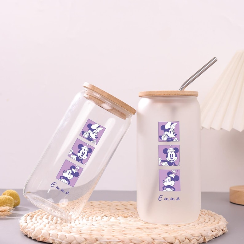Puede incluir: Dos vasos de vidrio con tapas de bamb&uacute; y pajitas de metal. Uno es transparente y el otro es esmerilado. Ambos presentan un gr&aacute;fico vertical morado con tres personajes de rat&oacute;n y el nombre Emma impreso en cursiva en la parte inferior.