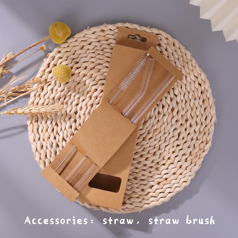 May include: A set of clear glass drinking straws and a thin cleaning brush packaged in a brown cardboard sleeve. The packaging features cutouts to display the items and includes the text Accessories: straw, straw brush printed at the bottom.