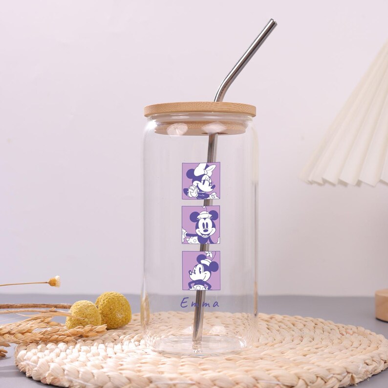 Puede incluir: Un vaso de cristal transparente con tapa de bamb&uacute; y pajita de metal curvada. El vaso presenta tres gr&aacute;ficos cuadrados morados apilados, cada uno con una ilustraci&oacute;n diferente de Minnie Mouse. El nombre Emma est&aacute; impreso en morado debajo.