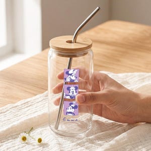 Puede incluir: Un vaso transparente con tapa de bamb&uacute; y pajita de metal curvada. El vaso presenta tres gr&aacute;ficos cuadrados morados apilados de un personaje de rat&oacute;n de dibujos animados con sombrero, con el nombre Emma impreso en letras cursivas moradas.