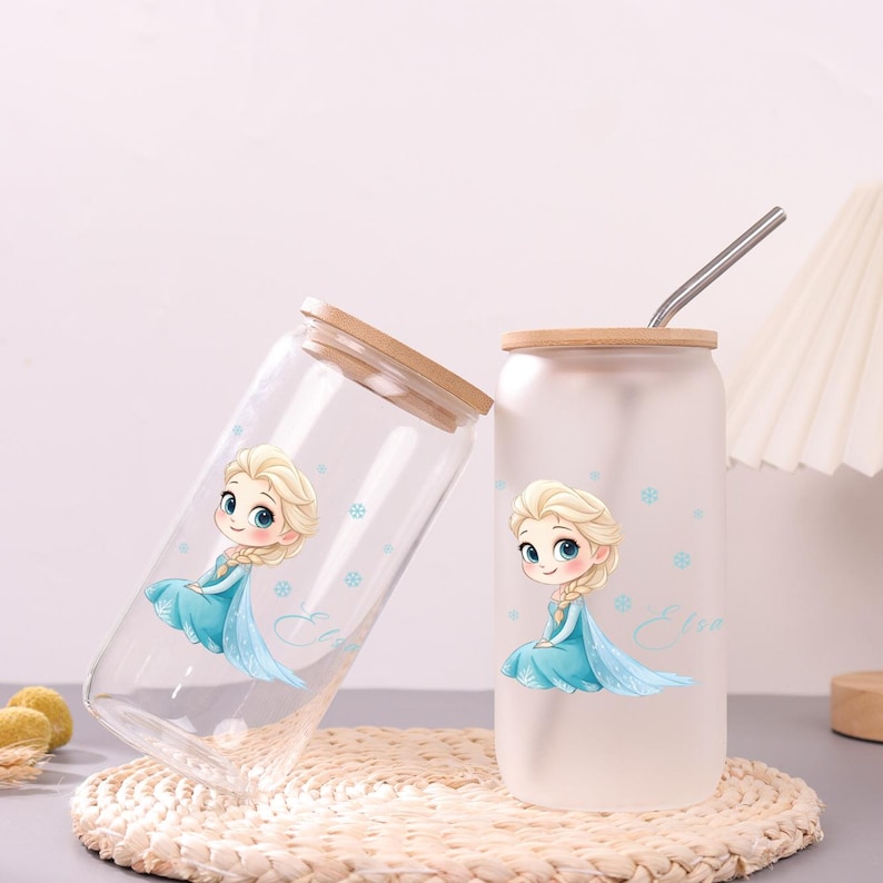 May include: Two glass tumblers with bamboo lids, one clear and one frosted, each featuring a cartoon illustration of a blonde character in a blue dress with snowflakes and the name Elsa written in cursive script, plus a metal straw.