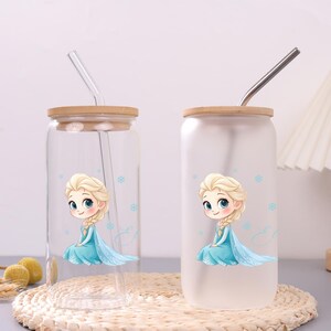 May include: Two glass tumblers with bamboo lids and straws. One tumbler is clear, the other is frosted. Both feature a cartoon illustration of a blonde character in a blue dress with light blue snowflakes and a cursive letter E design.
