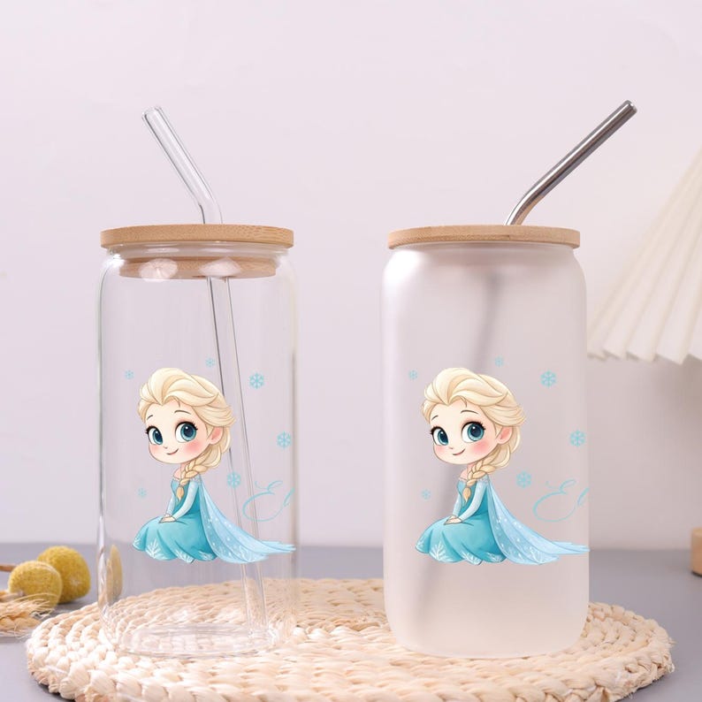 May include: Two glass tumblers with bamboo lids and straws. One tumbler is clear, the other is frosted. Both feature a cartoon illustration of a blonde character in a blue dress with light blue snowflakes and a cursive letter E design.