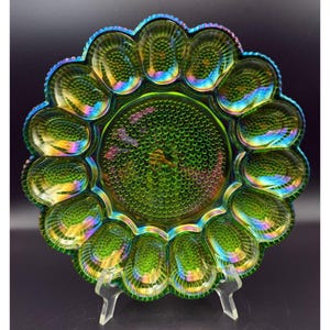 Può includere: Un piatto portauova rotondo in vetro iridescente verde caratterizzato da una sezione circolare centrale con trama a rilievo, circondata da dodici rientranze ovali individuali con bordi scanalati e un bordo esterno smerlato e perlato.