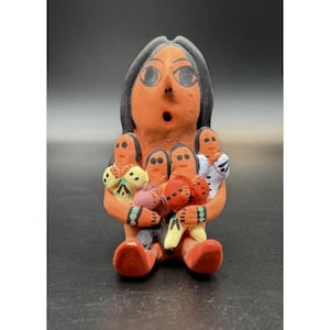 May include: A terracotta-coloured figurine of a woman holding several smaller figures. The woman has black hair and large eyes. The smaller figures have colourful clothing, including yellow, white, and red. The figurine is on a black surface.