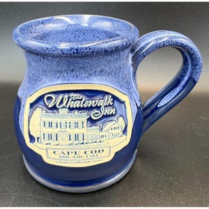 Könnte beinhalten: Blaue Keramiktasse mit Henkel. Die Tasse hat ein cremefarbenes rechteckiges Emblem mit den Worten "The Whalewalk Inn" und einer Illustration eines Gebäudes. Die Worte "Cape Cod" und eine Telefonnummer sind ebenfalls sichtbar.