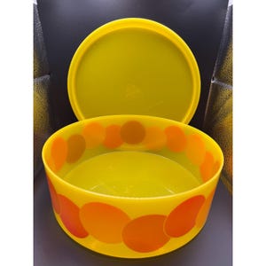 Puede incluir: Un cuenco de plástico amarillo vintage con tapa a juego. El cuenco está decorado con grandes círculos naranjas y amarillos alrededor del borde. La tapa está a un lado, revelando el interior del cuenco.