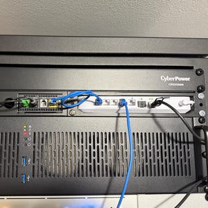 May include: A black server rack with multiple components, including a CyberPower unit, network devices, and various cables. The rack has handles and a red light indicator. Blue and white cables are connected to the devices.