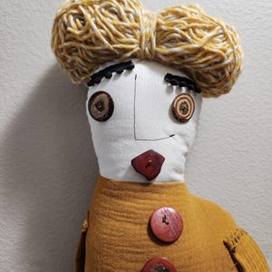 May include: A handmade doll with a white face, button eyes, and a wooden nose. The doll has a mustard-colored dress with large wooden buttons and a yarn hairstyle. The doll's features are stitched with black thread.
