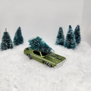 May include: A green toy car with a small green Christmas tree on top, surrounded by miniature snow-covered pine trees in a white snowy scene.