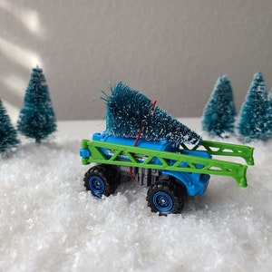 May include: A blue toy truck with a green frame hauling a small artificial Christmas tree. The truck is on a white surface that resembles snow.