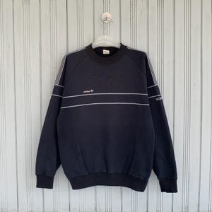Peut inclure: Un sweat-shirt bleu marine avec des rayures blanches et le logo Adidas. Le haut à manches longues a un col rond et des poignets côtelés. Le mot "TECH" est imprimé sur la manche.