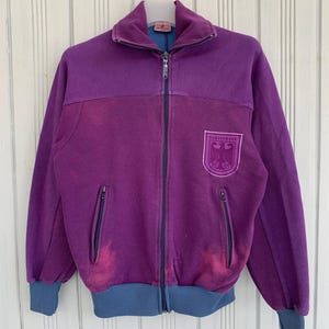May include: A vintage purple zip-up jacket with a high collar and two front pockets. The jacket features a blue collar, cuffs, and waistband. A crest with the text "BUNDESWEHR" is embroidered on the chest.