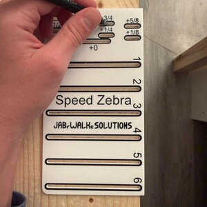 May include: A white Speed Zebra tool with black text and brown markings is held against a wooden surface. The tool has measurements and the text "JAB-WALK SOLUTIONS". A black marker is held above the tool.