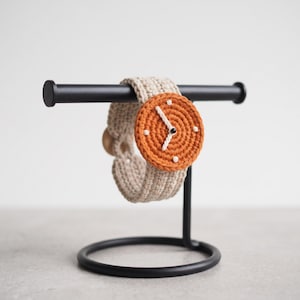 Puede incluir: Un reloj de ganchillo con una correa beige y una esfera circular naranja. La esfera presenta marcadores de hora blancos bordados y dos manecillas blancas centradas con una pequeña cuenta negra. El reloj se exhibe en un soporte de metal negro.