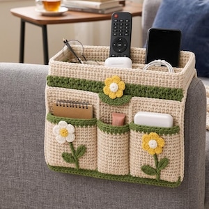 May include: A beige crochet sofa armrest organizer featuring three front pockets and one large back compartment. The organizer is decorated with green crochet trim and three yellow and white flower appliqués with green stems and leaves attached to the front.