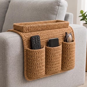 May include: A tan crochet sofa armrest organizer featuring a rectangular top tray and three vertical pockets. The pockets contain a black remote control, a black smartphone, and a pair of black-framed eyeglasses, providing storage for small items.