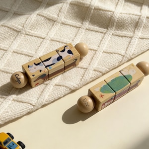 May include: Two wooden block puzzles with animal illustrations and wooden end pieces. One puzzle features a cow, the other a turtle. The name "Celya" is on one end piece, and "Eidan" on the other. A small yellow toy car is in the corner.