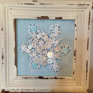 May include: A square, distressed white wooden frame containing a light blue felt background. Centered on the felt is a white snowflake cutout embellished with a dense arrangement of white buttons, clear beads, and small blue seed beads.