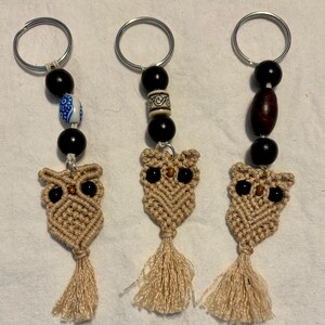 May include: Three macrame owl keychains featuring beige knotted cord bodies with tassel tails and black bead eyes. Each keychain includes a silver metal ring and a series of decorative beads, including solid black, patterned blue and white, or brown wood.