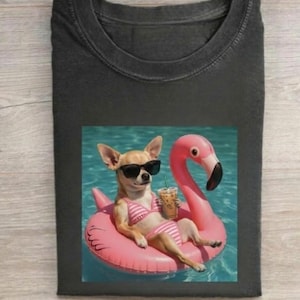 May include: Dark gray t-shirt featuring a chihuahua wearing sunglasses and a pink and white striped bikini, lounging on a pink flamingo float in a pool. The dog is holding a drink with a straw.