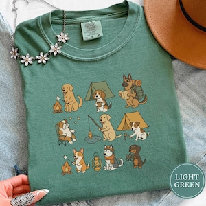 Puede incluir: Camiseta verde claro Comfort Colors con un gráfico de perros acampando. El diseño muestra varias razas de perros acampando, incluyendo asar malvaviscos, pescar y leer un mapa. La camiseta tiene cuello redondo y mangas cortas.