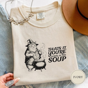 May include: Ivory-colored t-shirt with a graphic of a cat wearing a witch hat, stirring a cauldron. The text reads "THAT'S IT. YOU'RE GOING IN THE SOUP." A floral necklace and a brown hat are also visible.