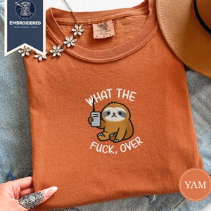 May include: Burnt orange t-shirt with a sloth graphic holding a walkie-talkie and the text "WHAT THE FUCK, OVER" in white. The shirt has a crew neck and short sleeves. The shirt is made of a soft, comfortable material.
