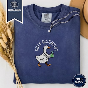 May include: A navy blue t-shirt with the embroidered phrase "Silly Scientist" above a cartoon goose wearing a lab coat and holding a green beaker. The shirt has a crew neck and short sleeves. A silver necklace with a star pendant is draped on the shirt.